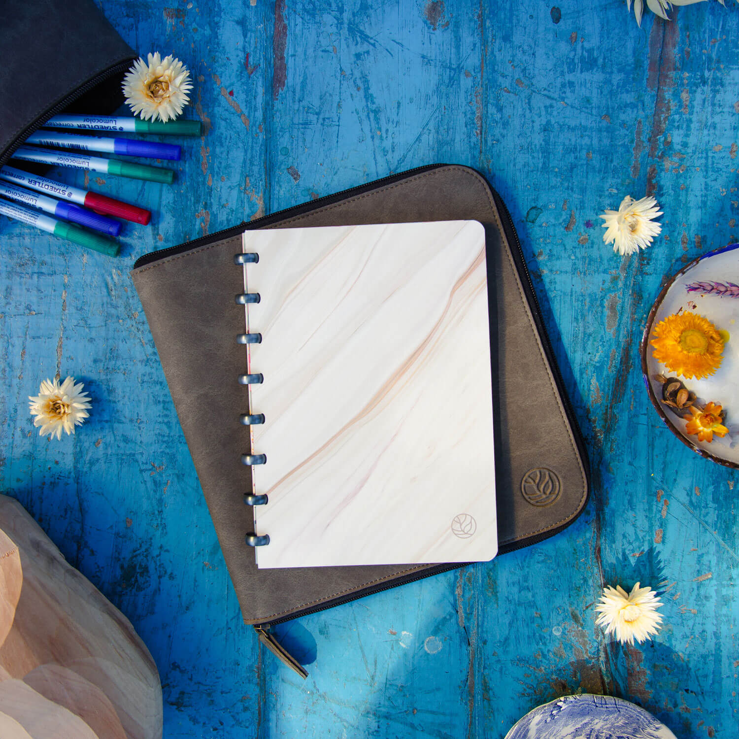 een uitwisbaar notitieboek met flowy marble kaft en spacegrey ringen en een clay werkmap, liggend op een antiek blauwe