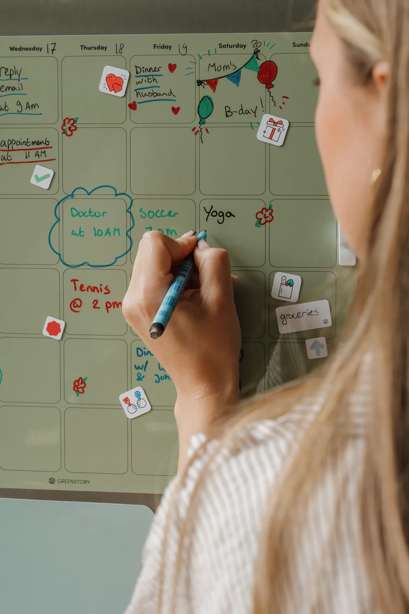 Vrouw schrijft een afspraak op een groene GreenStory weekplanner op de koelkast